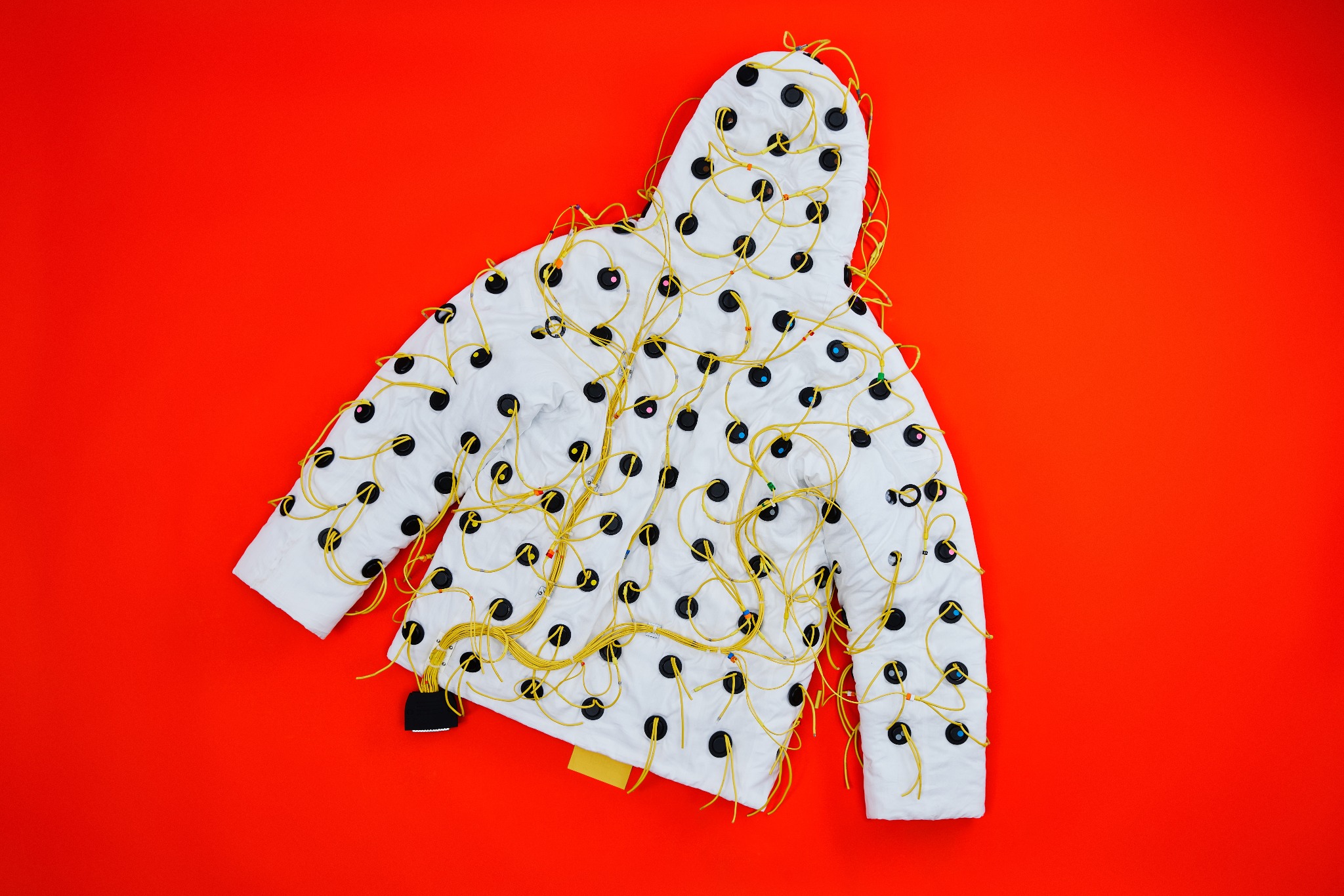 The white Sonic Jacket laid flat on a red background, with black speakers and yellow wires across its torso, sleeves and hood.